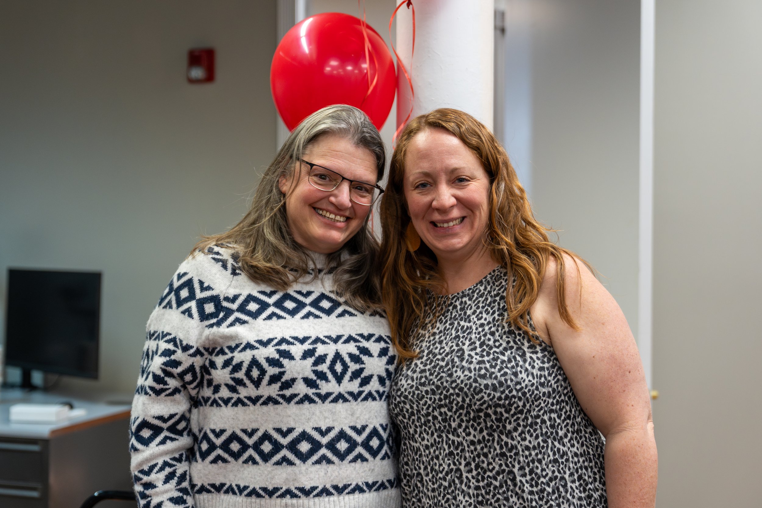 Two women smiling, one with glasses and a patterned sweater, and the other with curly red hair and a leopard print sleeveless top, standing close together with red balloons and streamers in the background.