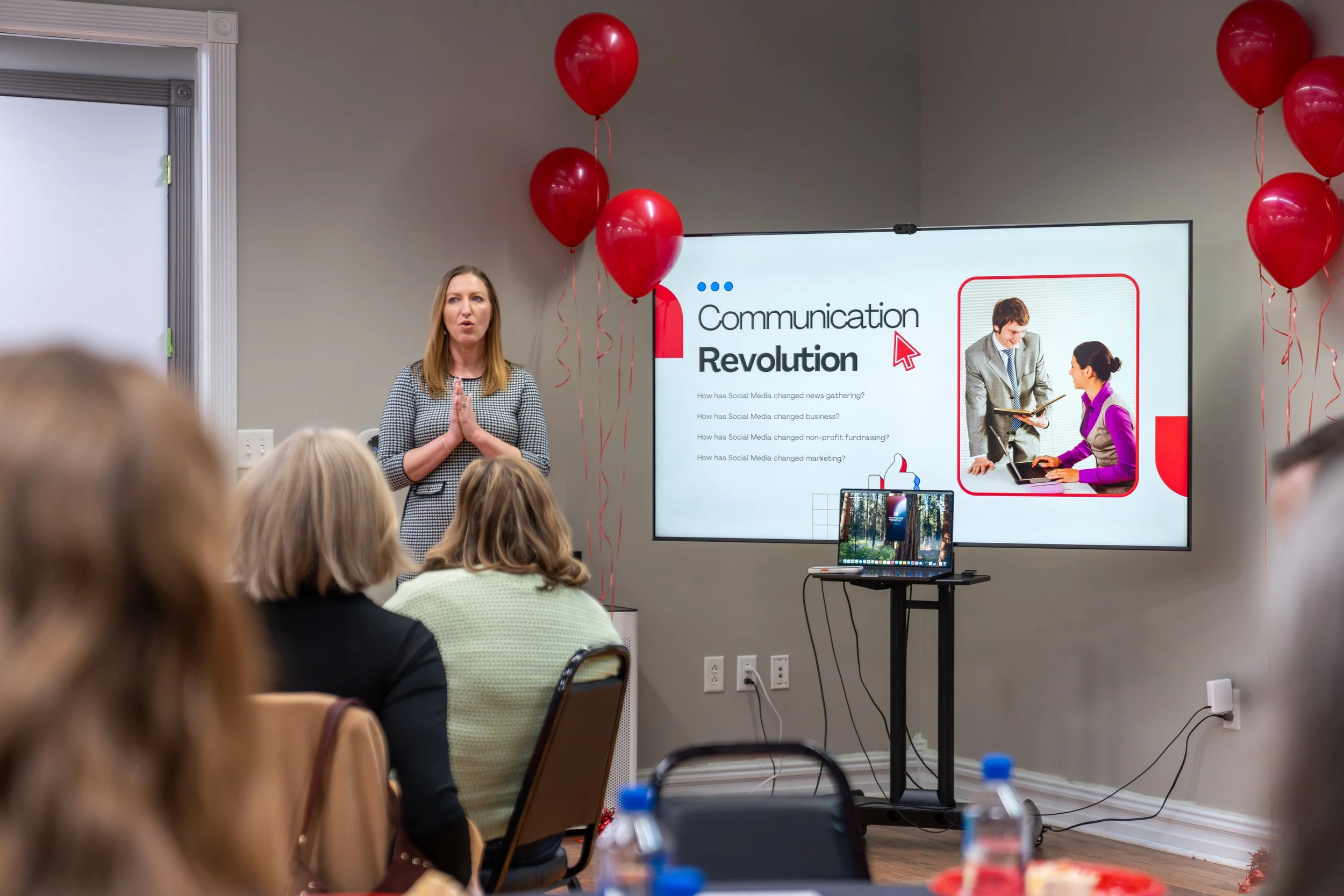 A woman giving a presentation to an audience in a conference room, with balloons hanging from the ceiling and a large screen displaying a slide titled 'Communication Revolution'.