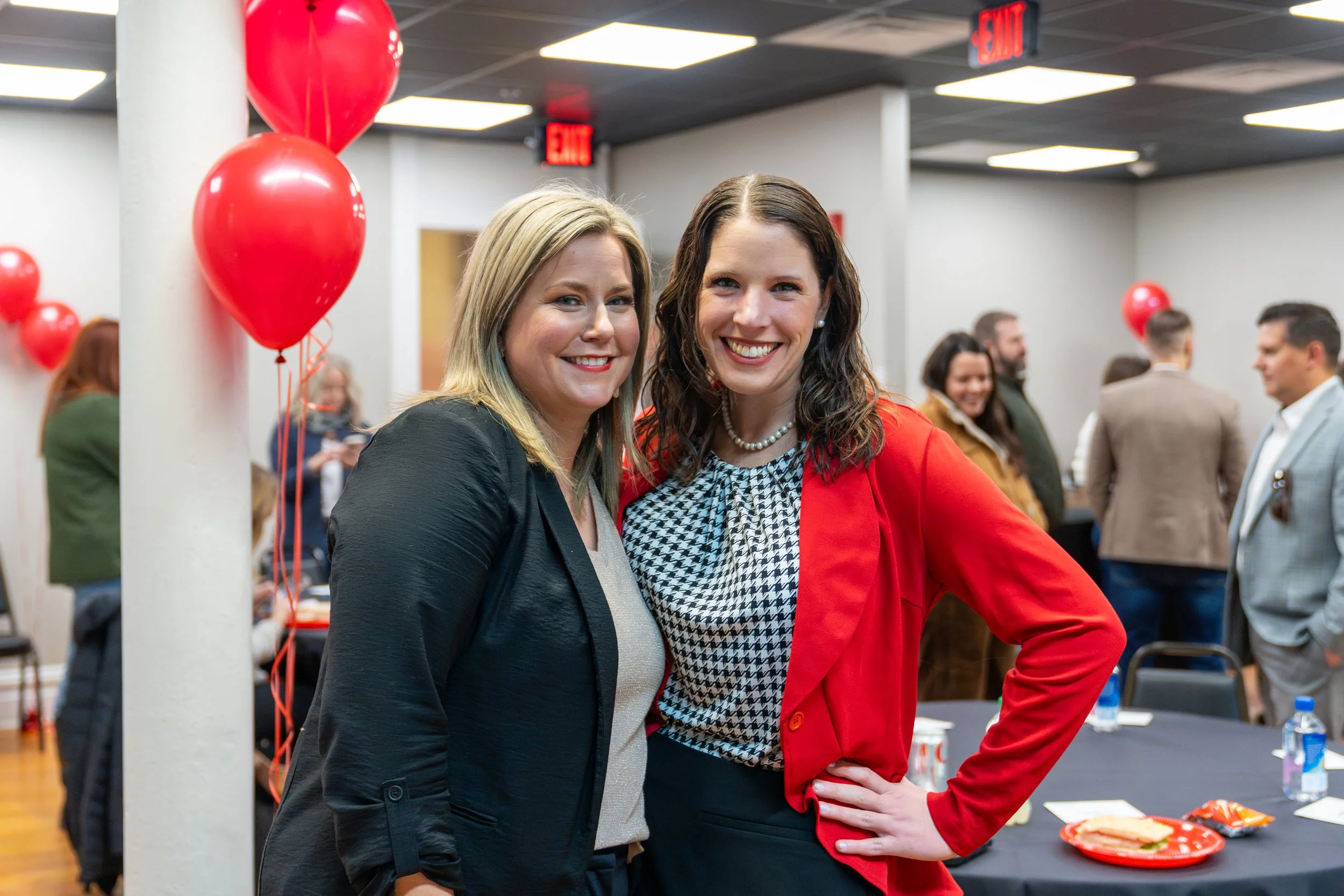 Two women smiling at the camera at a social gathering, with red balloons in the background.