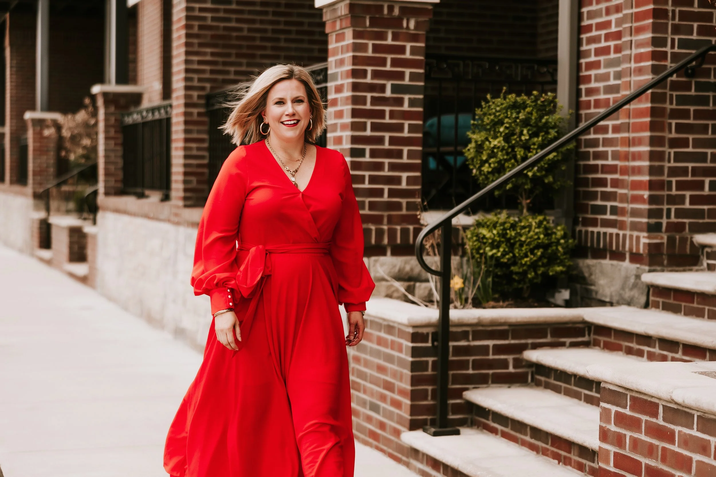 A woman wearing a bright red dress is smiling and walking along a sidewalk in front of a brick building with stairs and black railings.