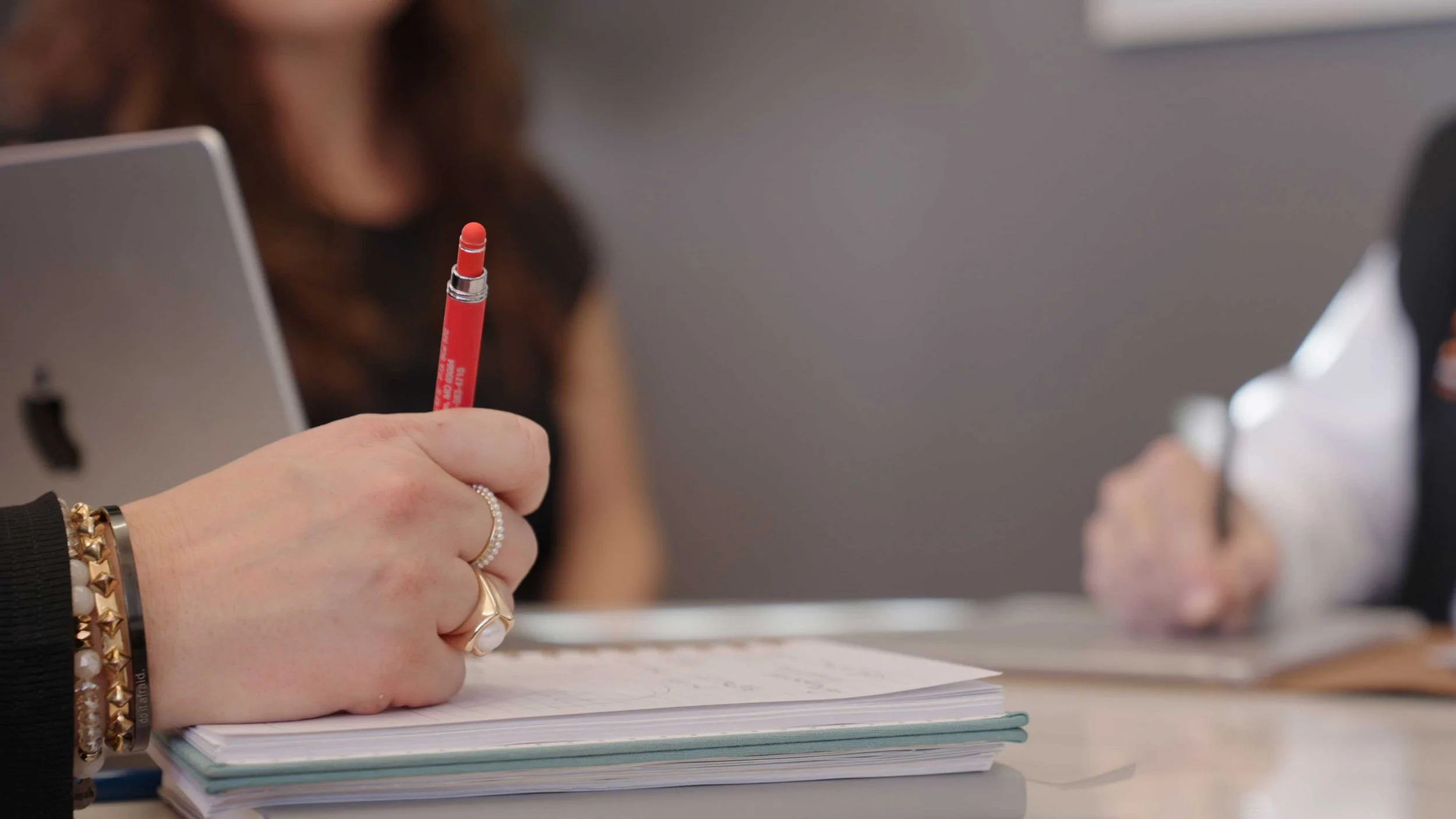 Close-up of a hand holding a red pen above a stack of papers on a table, with blurred individuals in the background.