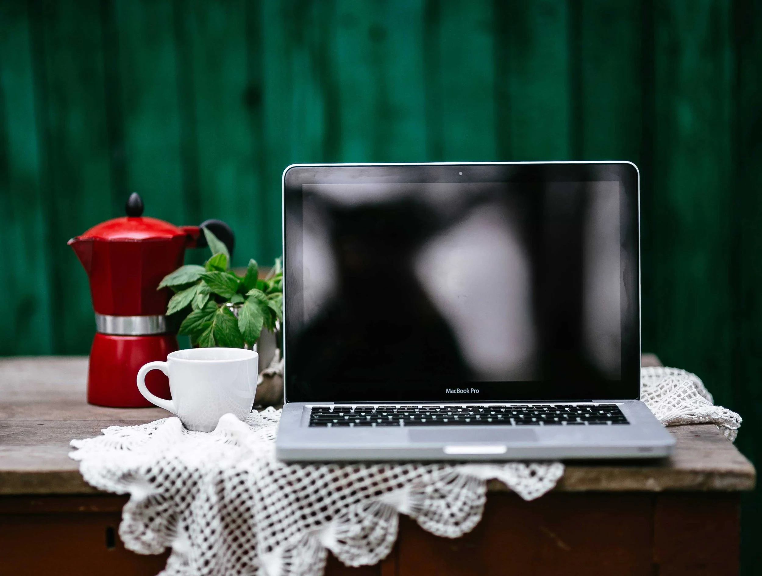 A MacBook Pro placed on a wooden table covered with a white crochet tablecloth, with a white mug, a red coffee pot, and a green potted plant nearby, set against a green background.