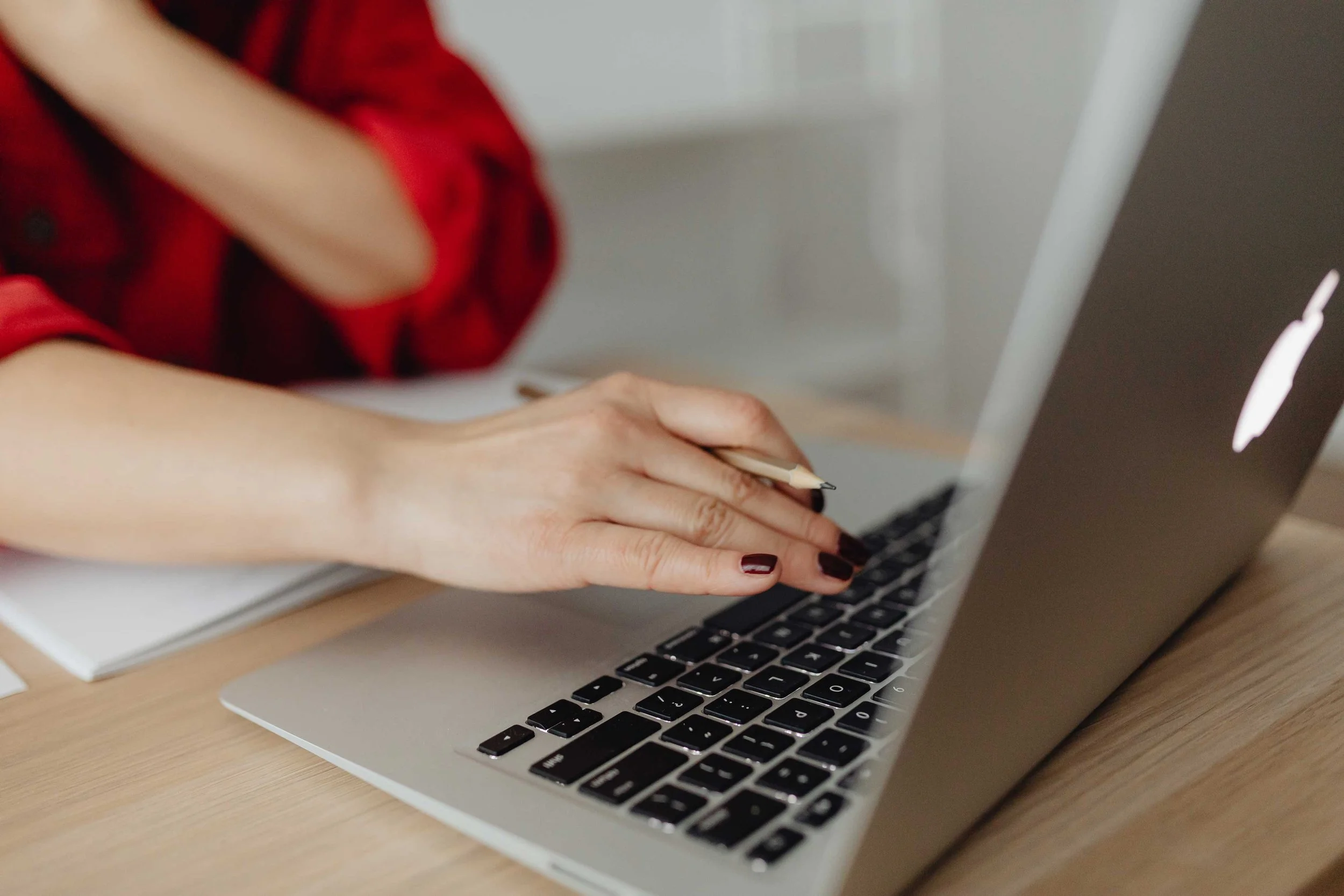 Close-up of a person's hand typing on a laptop keyboard, with a notebook and pen visible on a wooden desk.