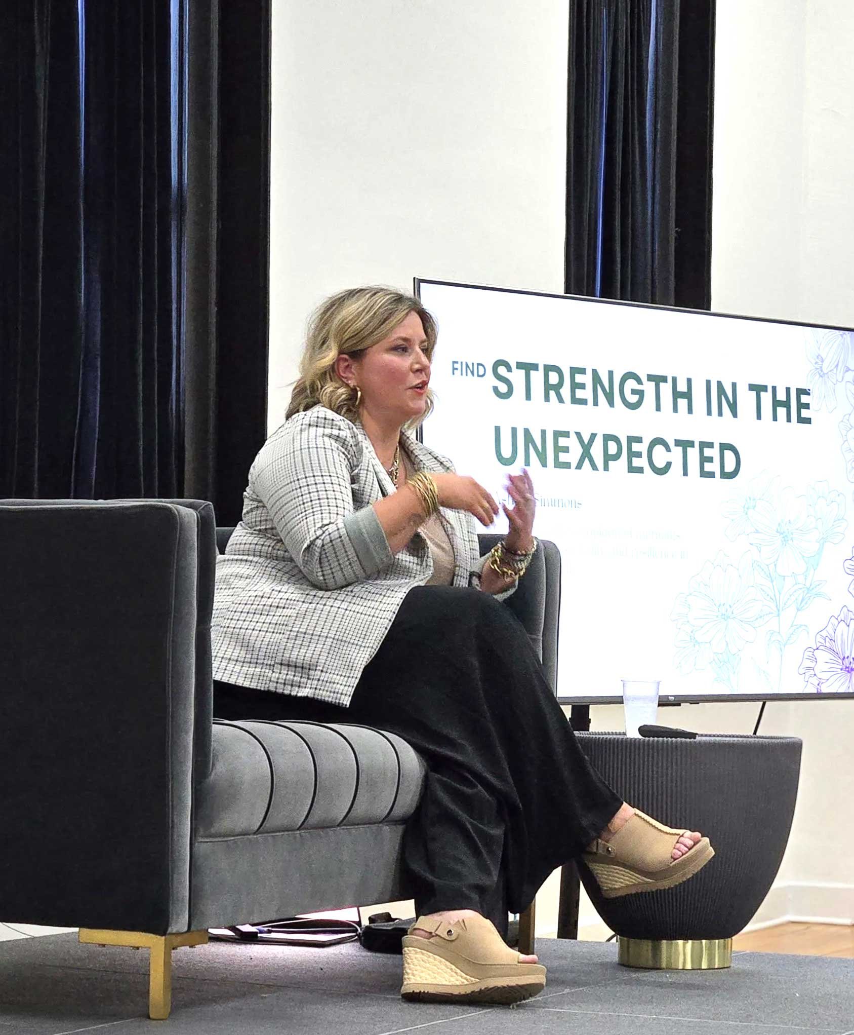 A woman sitting on a gray couch wearing beige platform sandals and a gray blazer, with a large screen behind her displaying text that reads 'Find Strength in the Unexpected'.