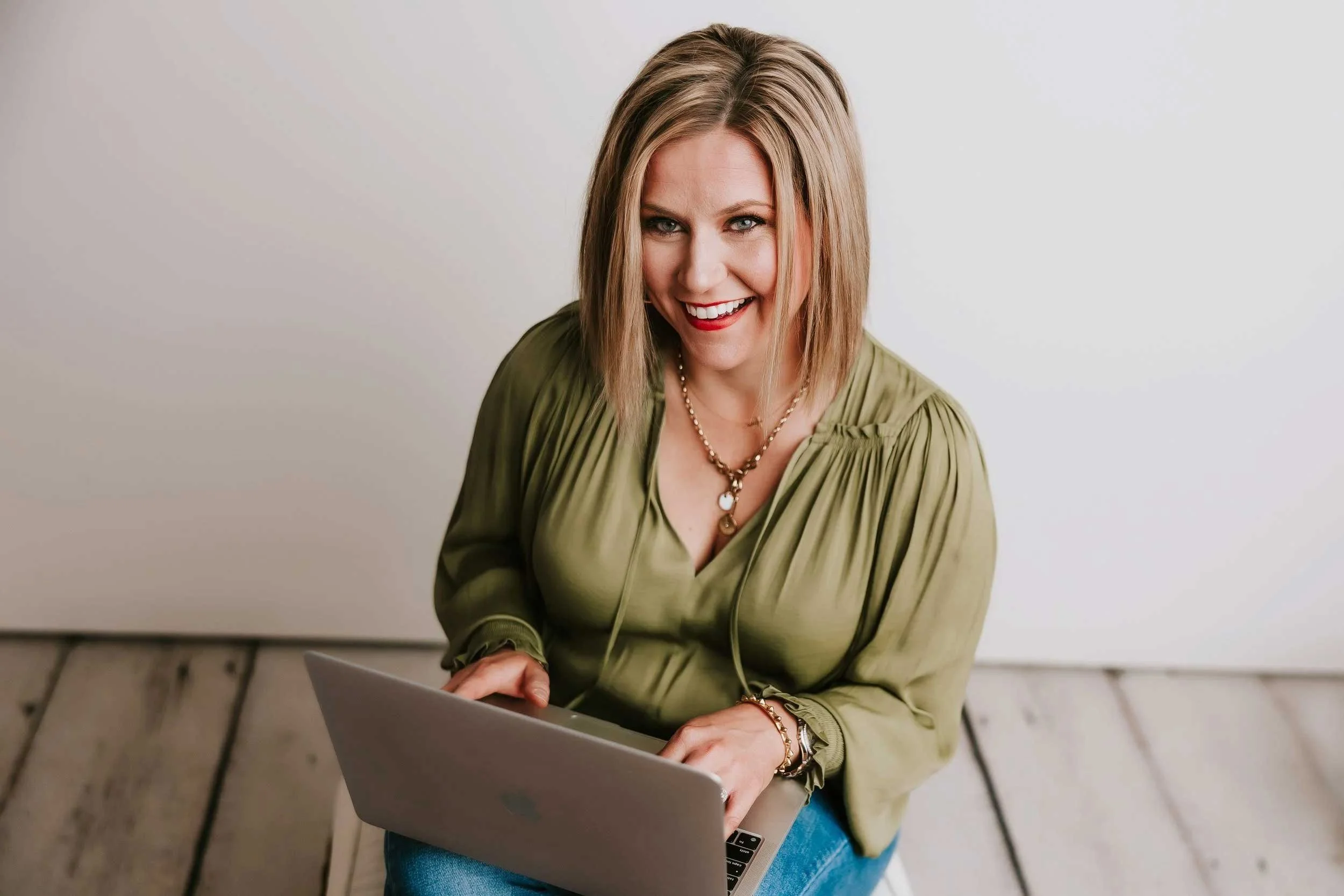 A woman with shoulder-length blonde hair, wearing a light green blouse and a pearl necklace, smiling while working on a laptop.