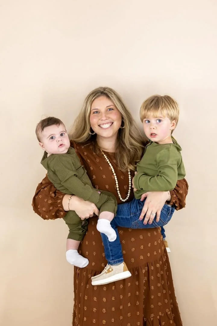 A woman with long blond hair, wearing a brown dress with a pattern and a string of pearls, holding a toddler in her left arm and a young boy in her right arm, both children wearing green hoodies. The woman is smiling broadly, while the children have surprised expressions. The background is plain beige.