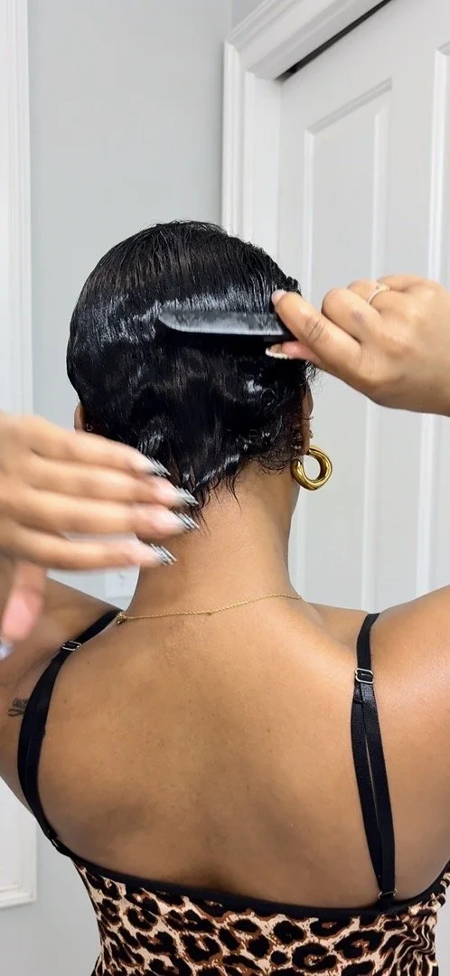 Person with short black hair using a brush to comb their wet hair in front of a white door, wearing a black strap top with a leopard print pattern and gold hoop earrings.