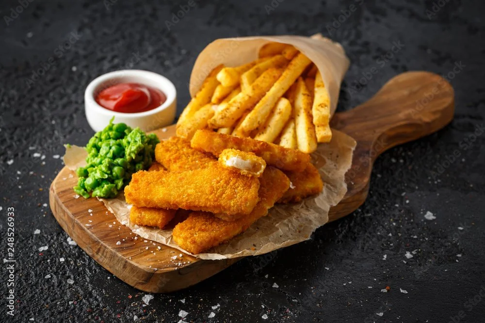 Fish and chips served on a wooden board with fried fish fillets, French fries, green peas, and a small bowl of ketchup.