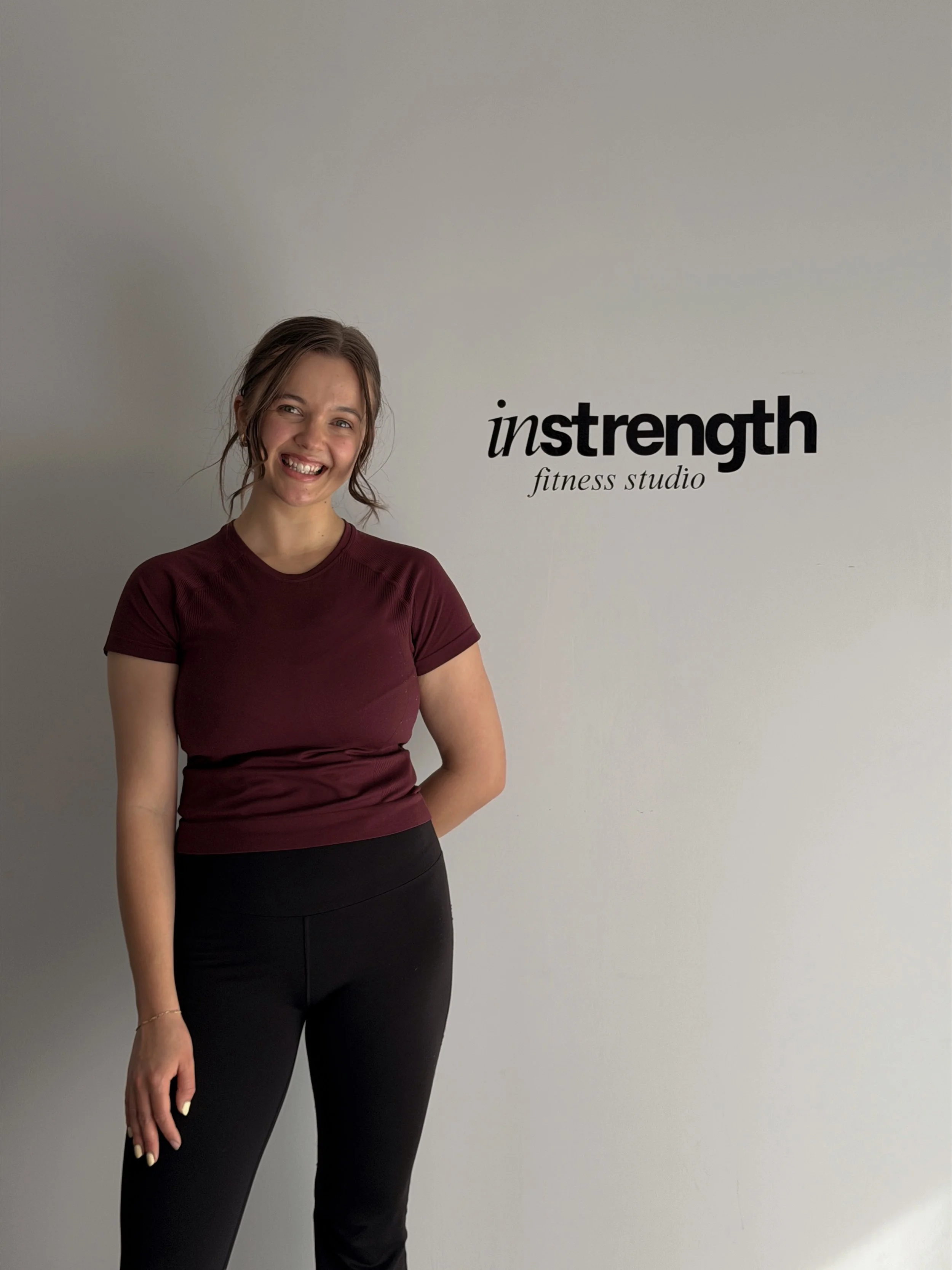 A smiling woman standing in front of a white wall with black text that reads "instretch fitness studio." She is wearing a maroon t-shirt and black pants.