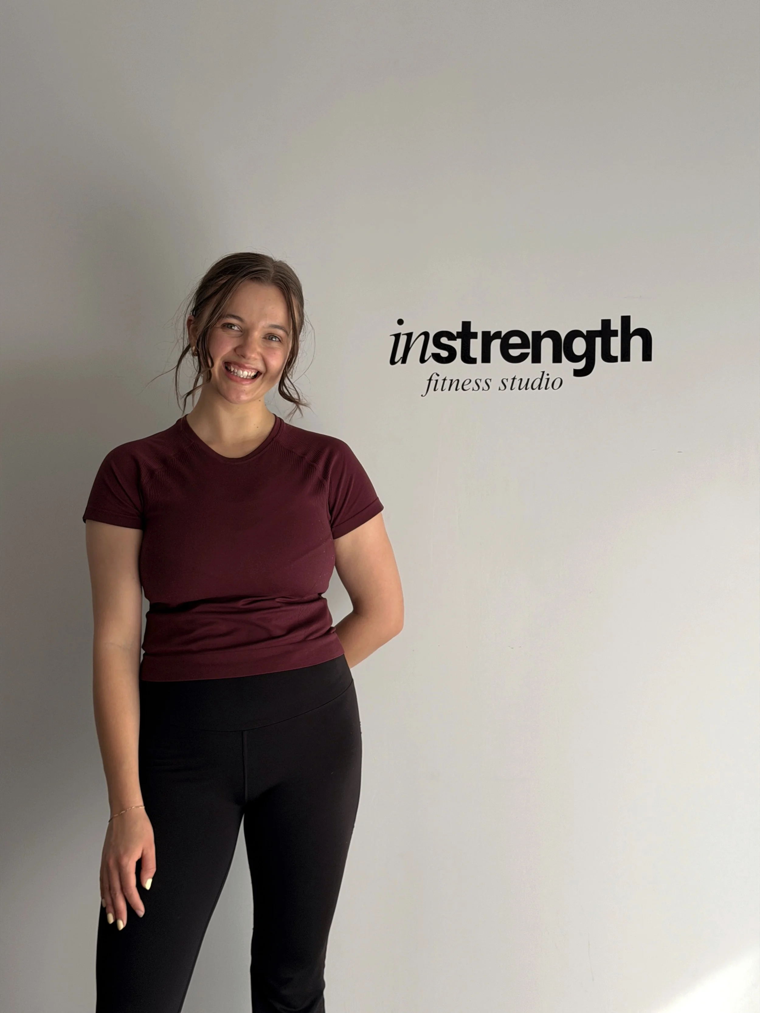 A woman with brown hair in a ponytail, wearing a maroon athletic shirt and black leggings, standing in front of a white wall with the words 'instrength fitness studio' in black text.