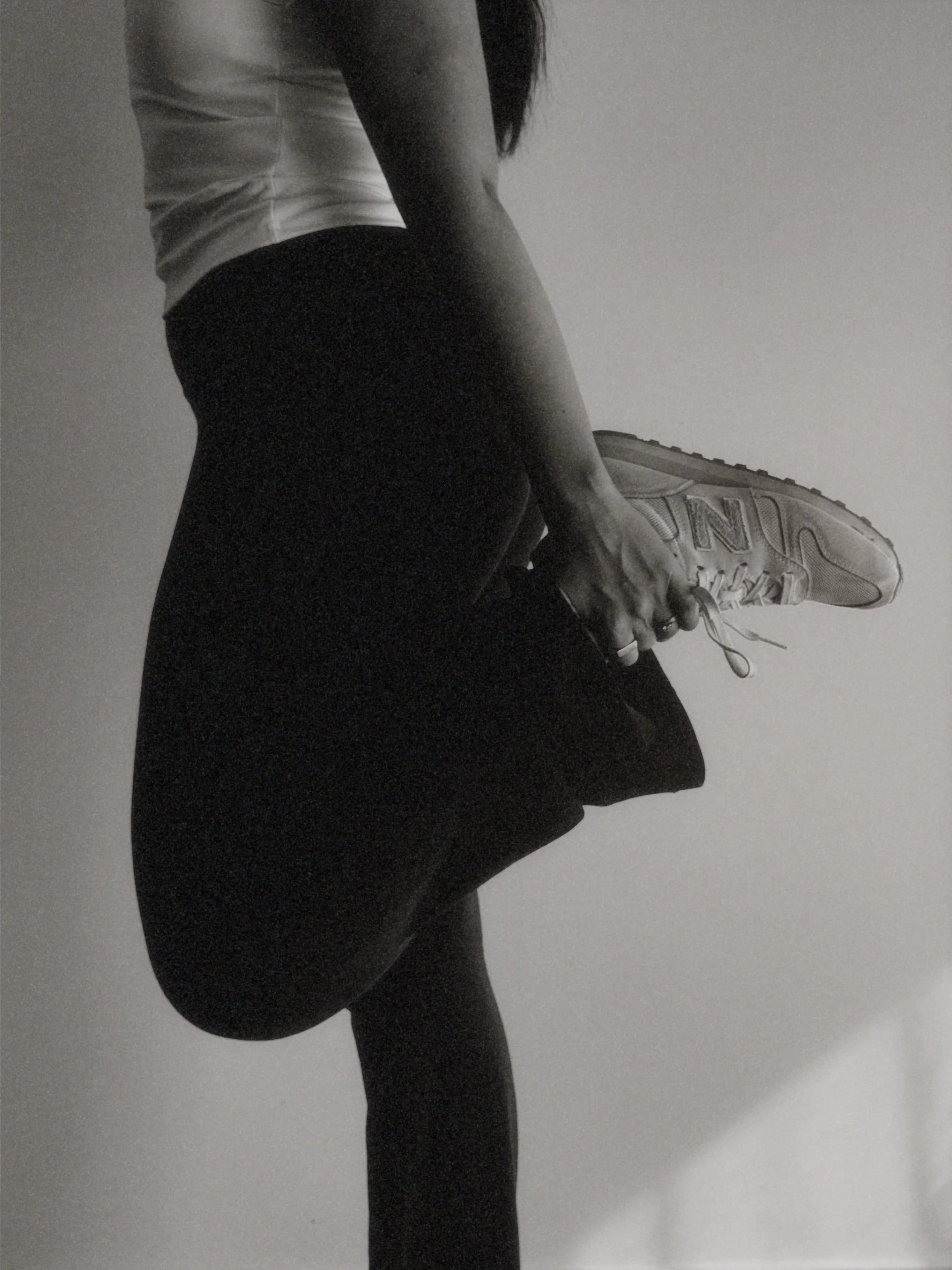 A person in athletic clothing tying their shoelace, with a focus on their legs and torso, against a plain wall background.