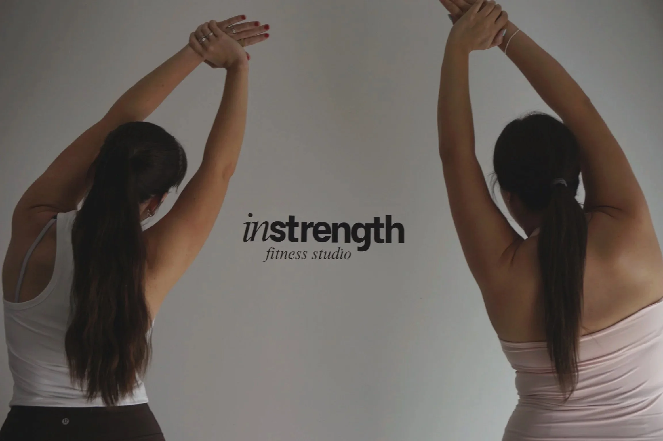 Two women are stretching with their arms raised above their heads, standing in front of a wall with the text "instrength fitness studio." Both women have long dark hair, and are dressed in athletic wear, one in a white top and the other in a striped top.