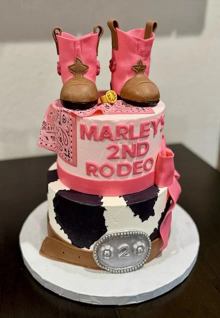 Tall pink and brown cowboy boots cake topper, sitting on a pink band with the words "Marley's 2nd Rodeo" in pink letters. The cake has a cow print design, a silver buckle with the number "2" inside, and a pink ribbon decoration.