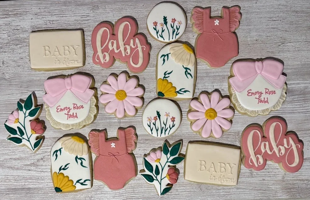 Collection of decorated cookies with baby shower themes, including cookies shaped like baby clothes, flowers, and words like "baby" and "Emory Rose Todd" on a wooden surface.
