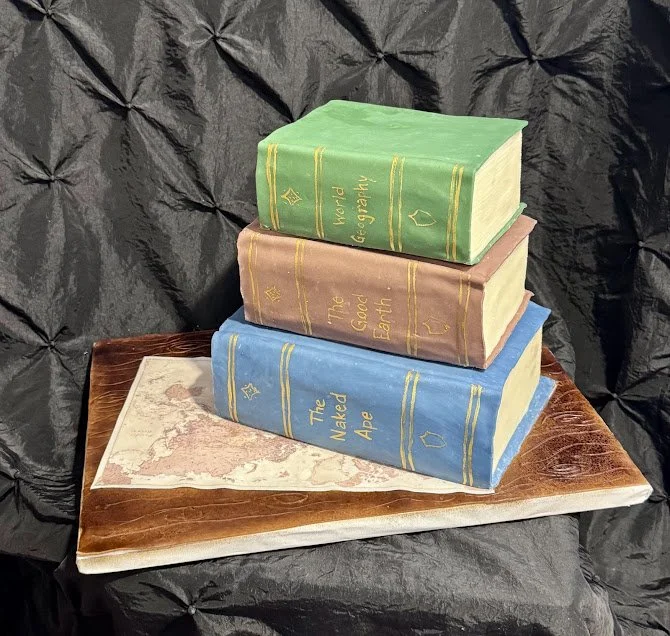 Cake shaped like three stacked books titled 'The Naked Ape', 'The Good Earth', and 'World Geography', on a base resembling an open map.