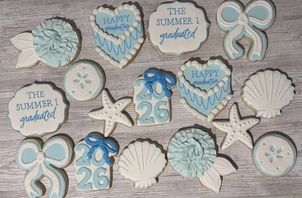 Cookies shaped like seastars, bows, shells, and message plaques in blue and white, with some decorated with icing and text celebrating graduation and summer, on a wooden surface.
