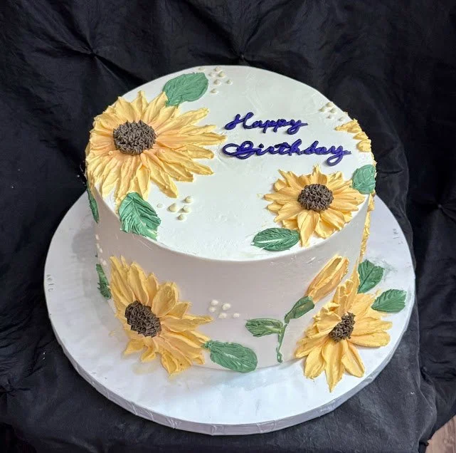 Round birthday cake decorated with large yellow sunflower icing flowers and green leaves, with a message in blue icing that says "Happy Birthday" on top.