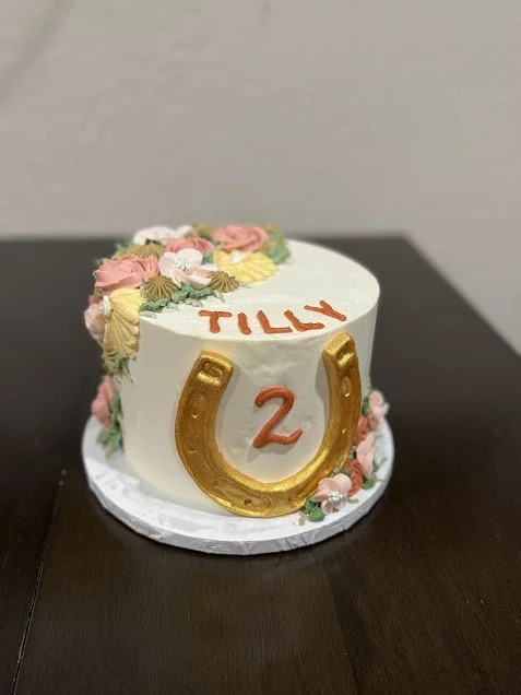 A round white birthday cake decorated with pink, yellow, and green floral frosting, with the name 'Tilly' written in pink, a number '2' in the center, and a large gold horseshoe on the side, sitting on a white cake board.