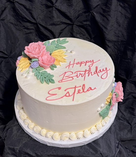 White birthday cake with floral decorations and pink writing that says "Happy Birthday Estela" on top.