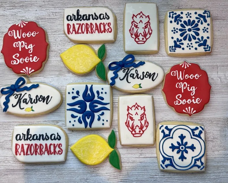 Decorative cookies with Arkansas Razorbacks, 'Woo Pig Sooie' slogans, blue floral patterns, a lemon, and a wild hog face logo, arranged on a light wooden surface.