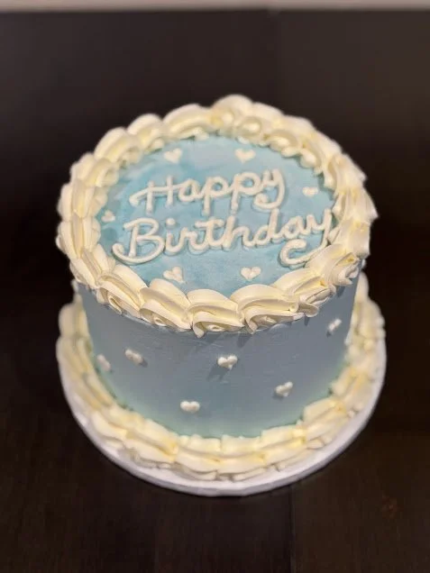 A round birthday cake with light blue frosting, white decorative hearts, and white frosting piping around the top and bottom edges, with "Happy Birthday" written on top in white icing.