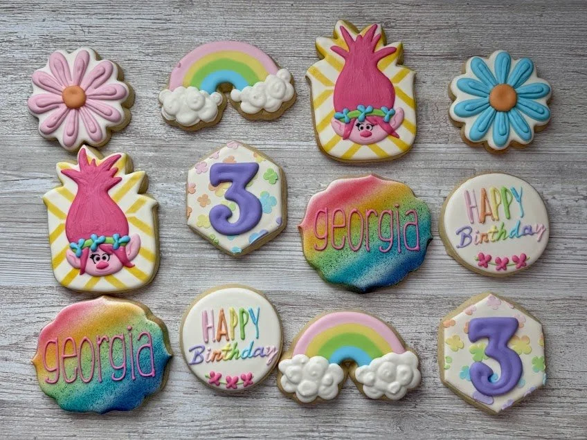 Trolls-themed decorated cookies featuring Poppy, rainbows with clouds, colorful rainbow-background cookies with the name "Georgia" and the number "3," and birthday messages.