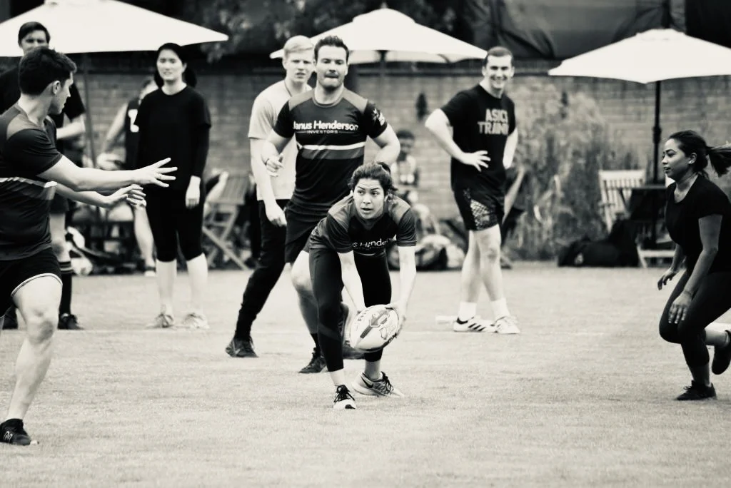 A group of people playing rugby outdoors on the grass field, with some holding a rugby ball and others observing, under large umbrellas.