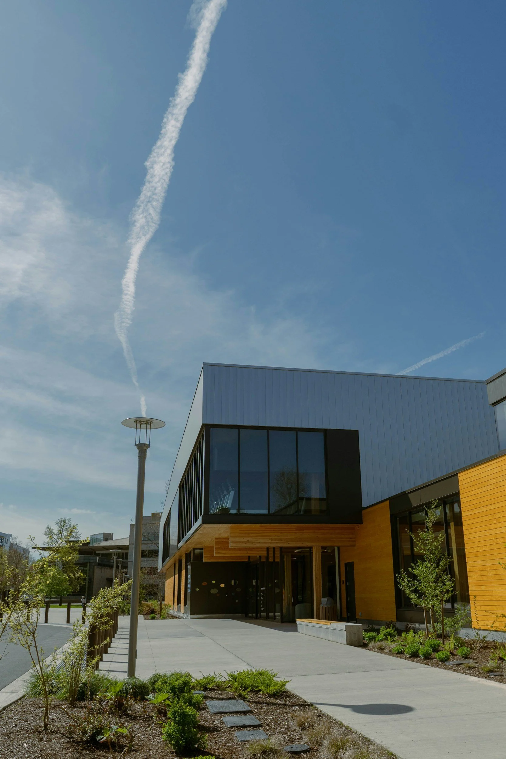 Modern building with large glass windows and wooden exterior, trees and bushes in landscaped area, cloudy sky with contrails. Street lamp nearby.