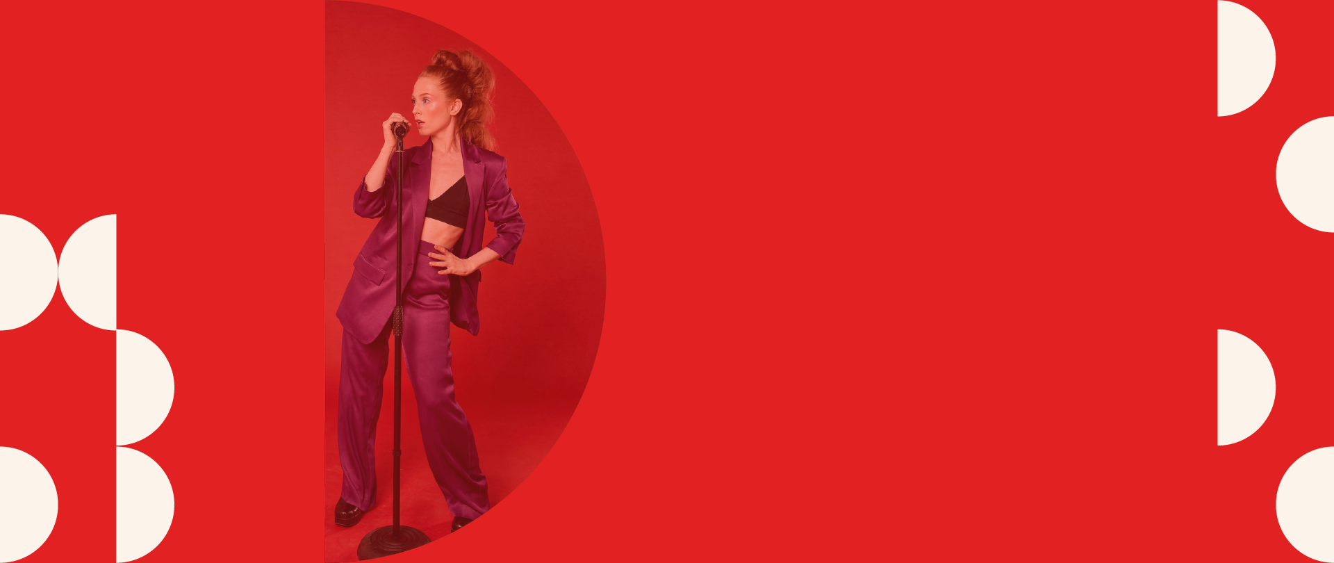 A woman in a purple suit and black crop top standing with one hand on her hip, holding a microphone in the other, against a red background with decorative white circular patterns.