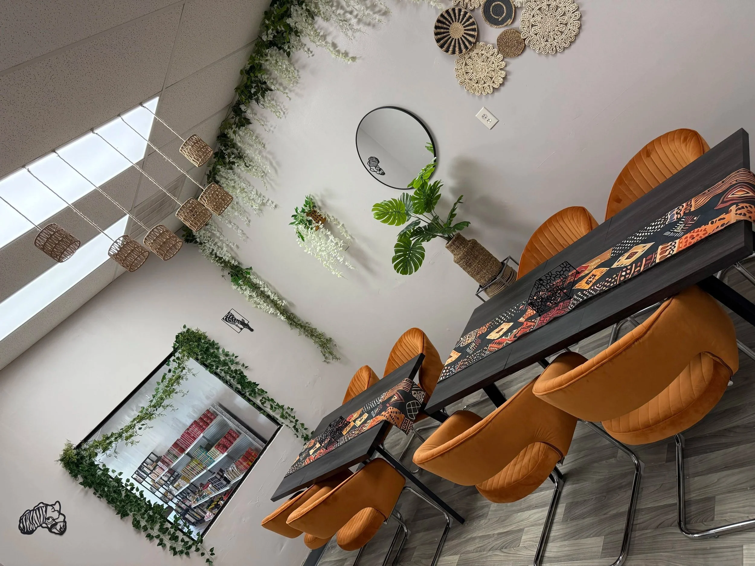 A modern dining room with a black table set with patterned table runner and orange chairs. Decor includes a round mirror, potted plant, and wall art. A wall with hanging lights and greenery accents is visible.