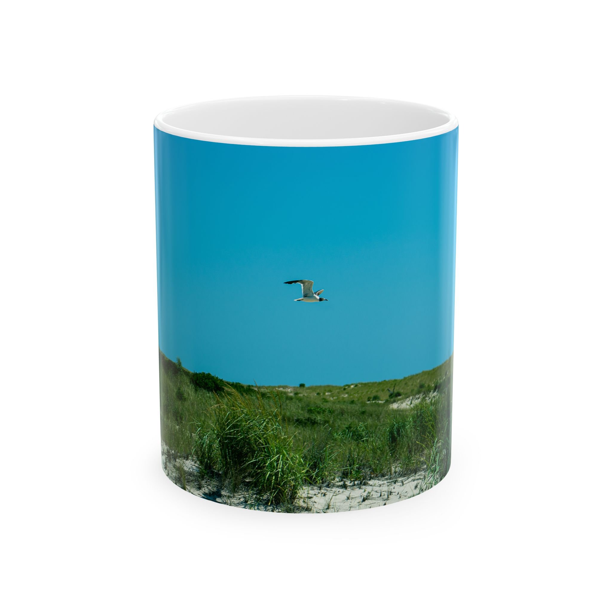 Island Speaks 'The Solitary Gull' 11 oz mug product photo. Front of mug shown that has photo of gull flying over some grassy dunes with the blue sky in the background.