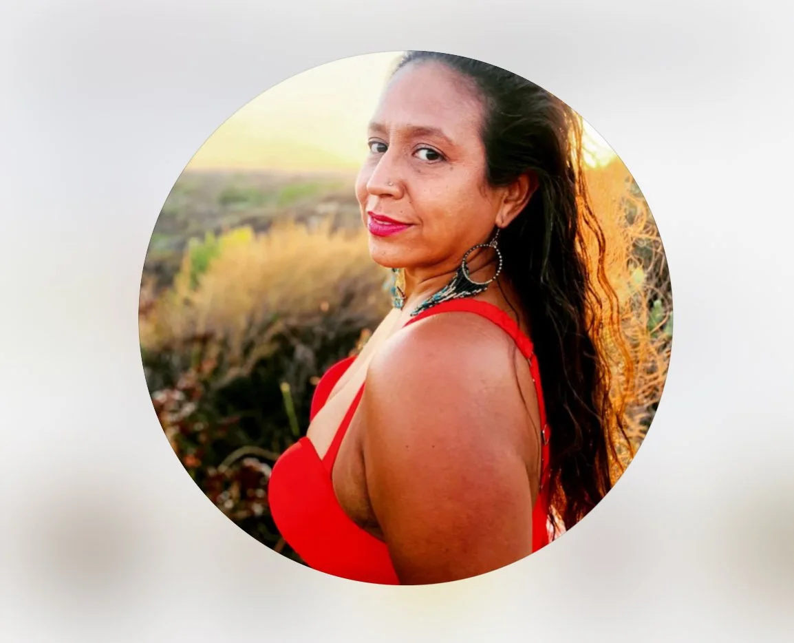 A woman with long dark hair wearing a red dress and hoop earrings, standing outdoors during sunset.