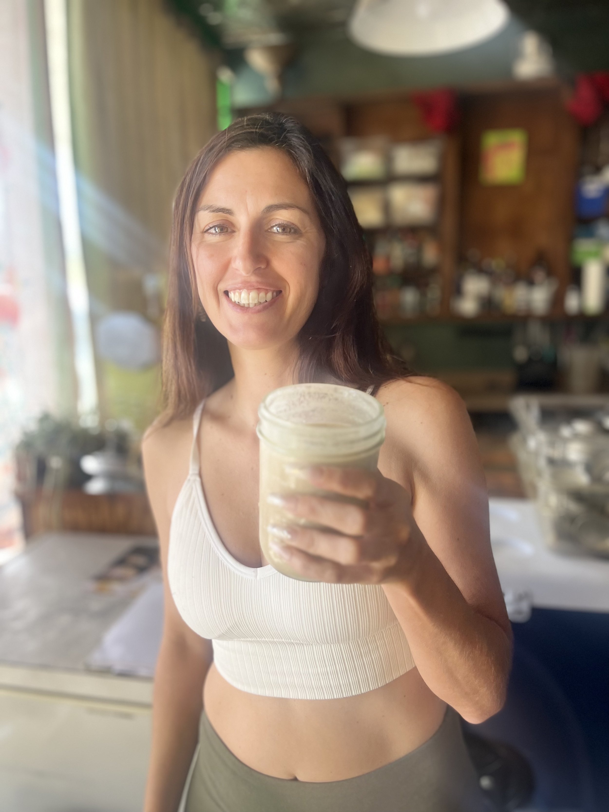 A woman smiling and holding a cup, standing in a room with shelves and a curtain in the background.