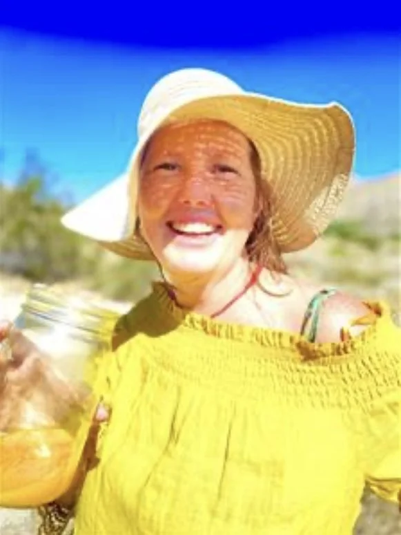 Woman in a yellow off-shoulder top and wide-brimmed straw hat smiling outdoors, holding a jar of yellow liquid.