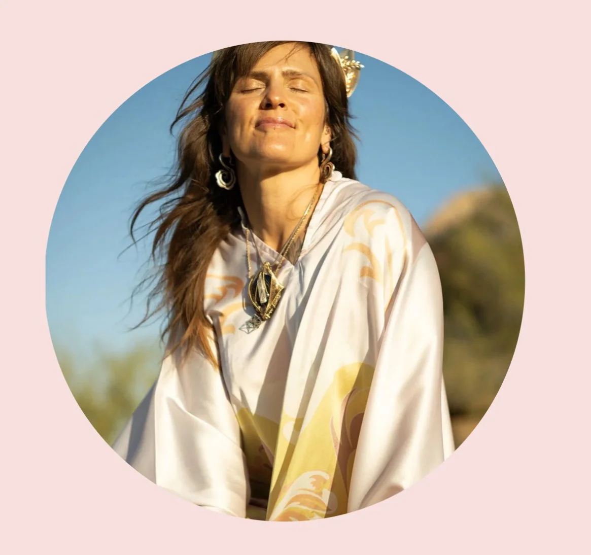 Woman with long brown hair wearing earrings and necklaces, sitting outdoors with eyes closed, smiling, in a white and yellow patterned dress, with a blue sky and mountains in the background.