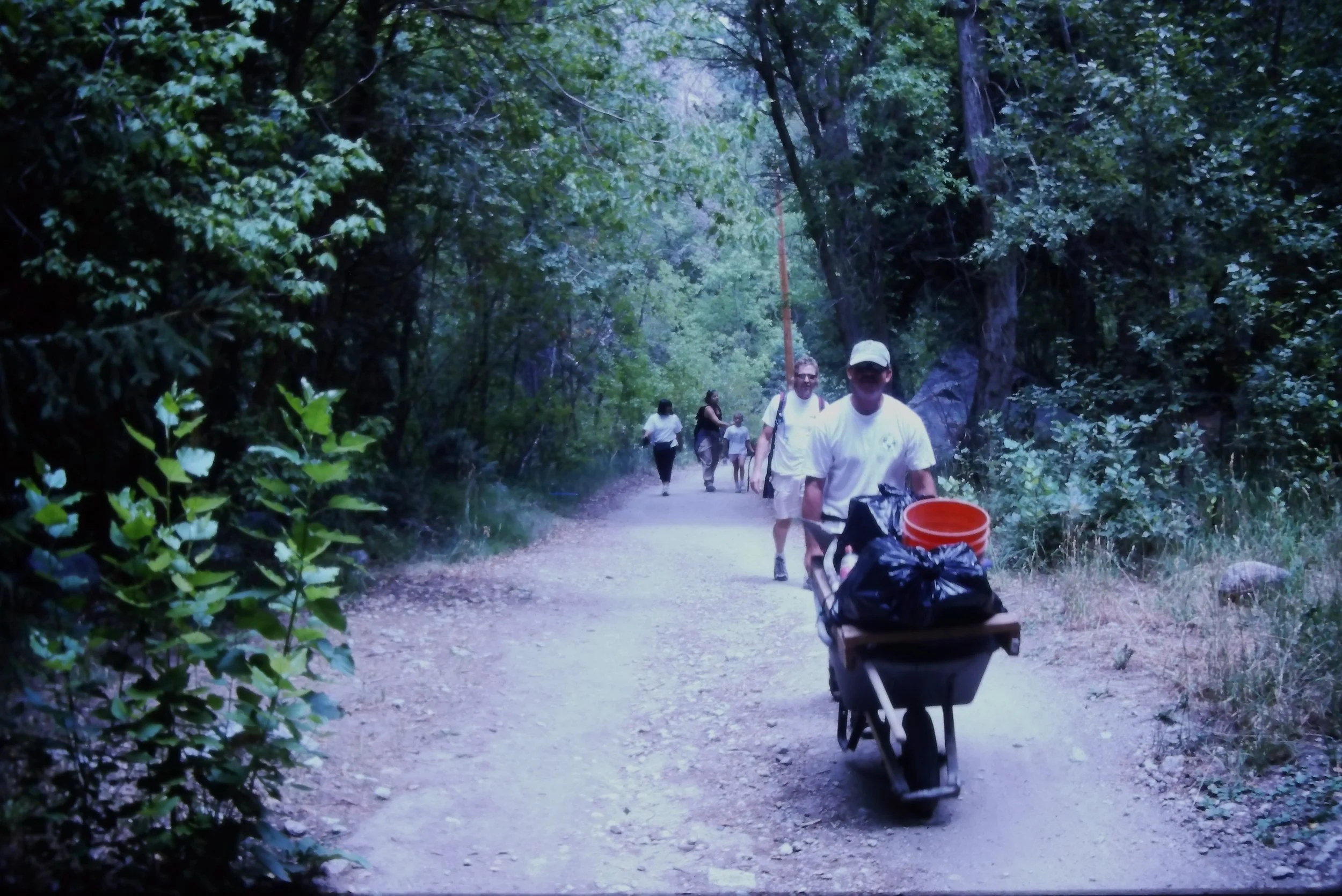 A group of people walking on a forest trail for an annual cleanup, with some pushing a wheelbarrow, surrounded by green trees and shrubs.