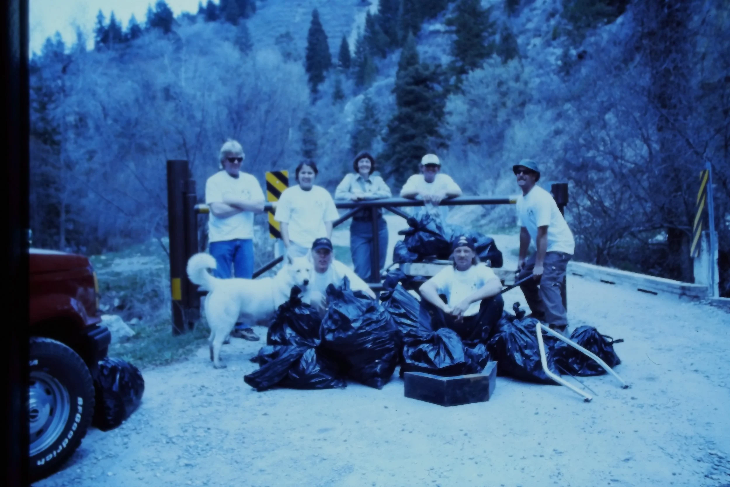 A group of people posing outdoors near the entrance to a canyon after annual cleanup in American Fork Canyon, Utah. There are bags and equipment on the ground, and a large dog is present in the foreground.