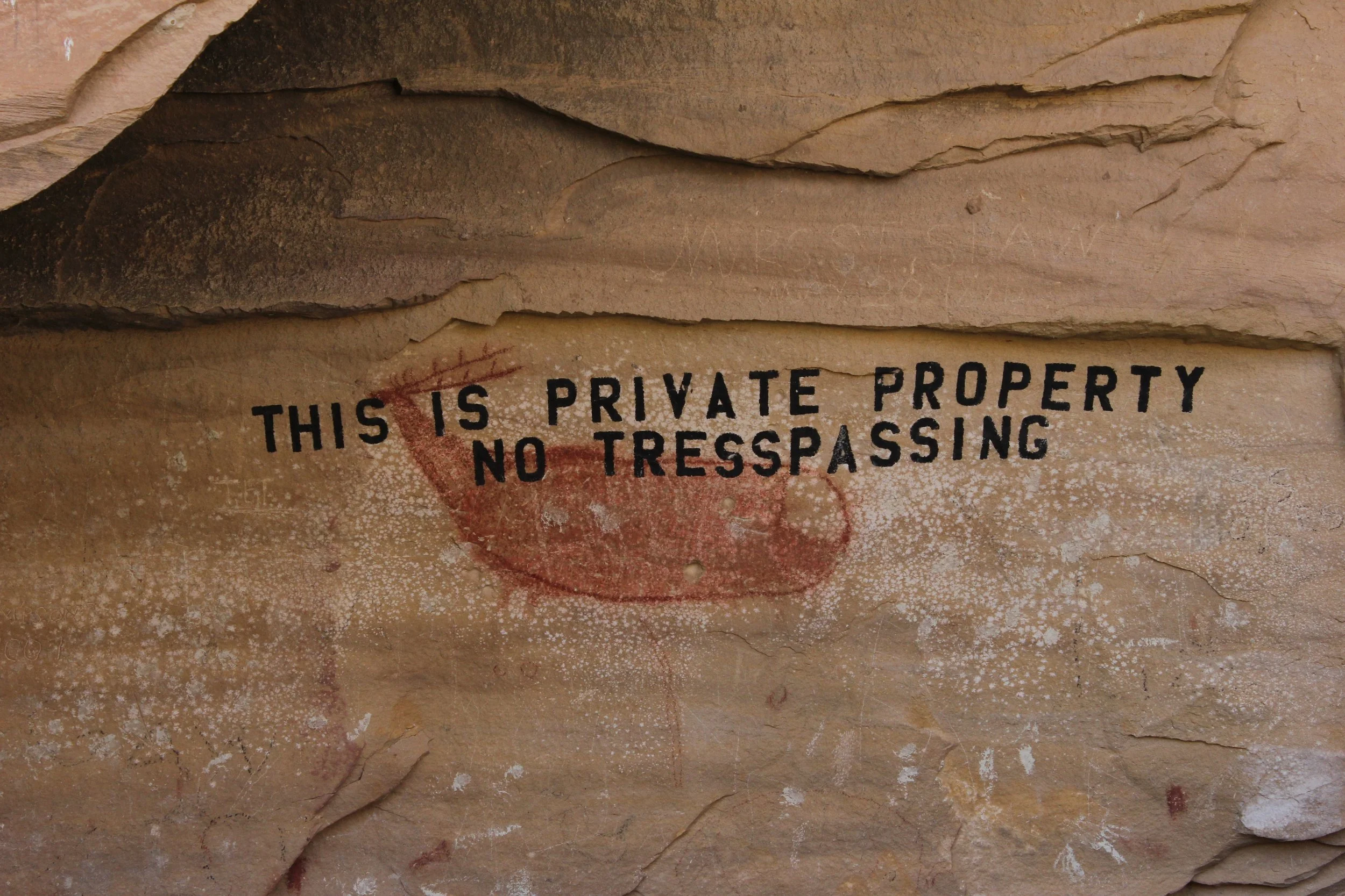 Close-up of a rock surface with the text 'This is private property no trespassing' carelessly painted over an ancient pictograph of a deer painted in red.