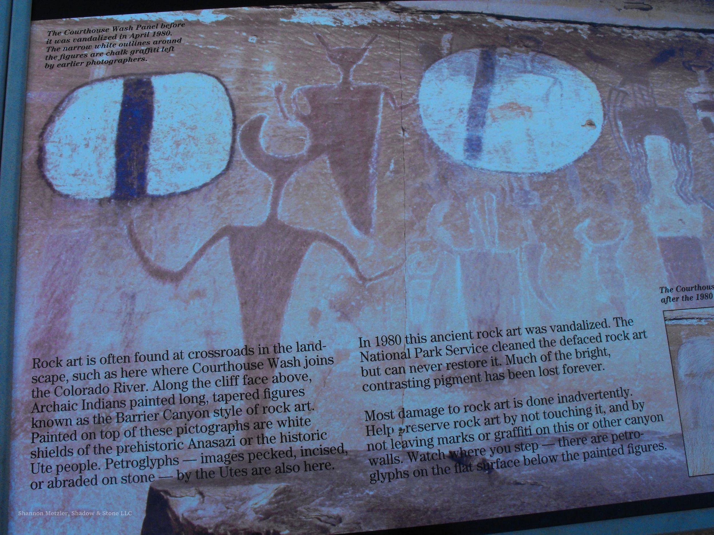 Photograph of an informational sign about graffiti vandalism on a rock art panel at a historic site. The sign includes a faint chalk graffiti left by earlier photographers, depicting two white painted figures and a central brown figure.