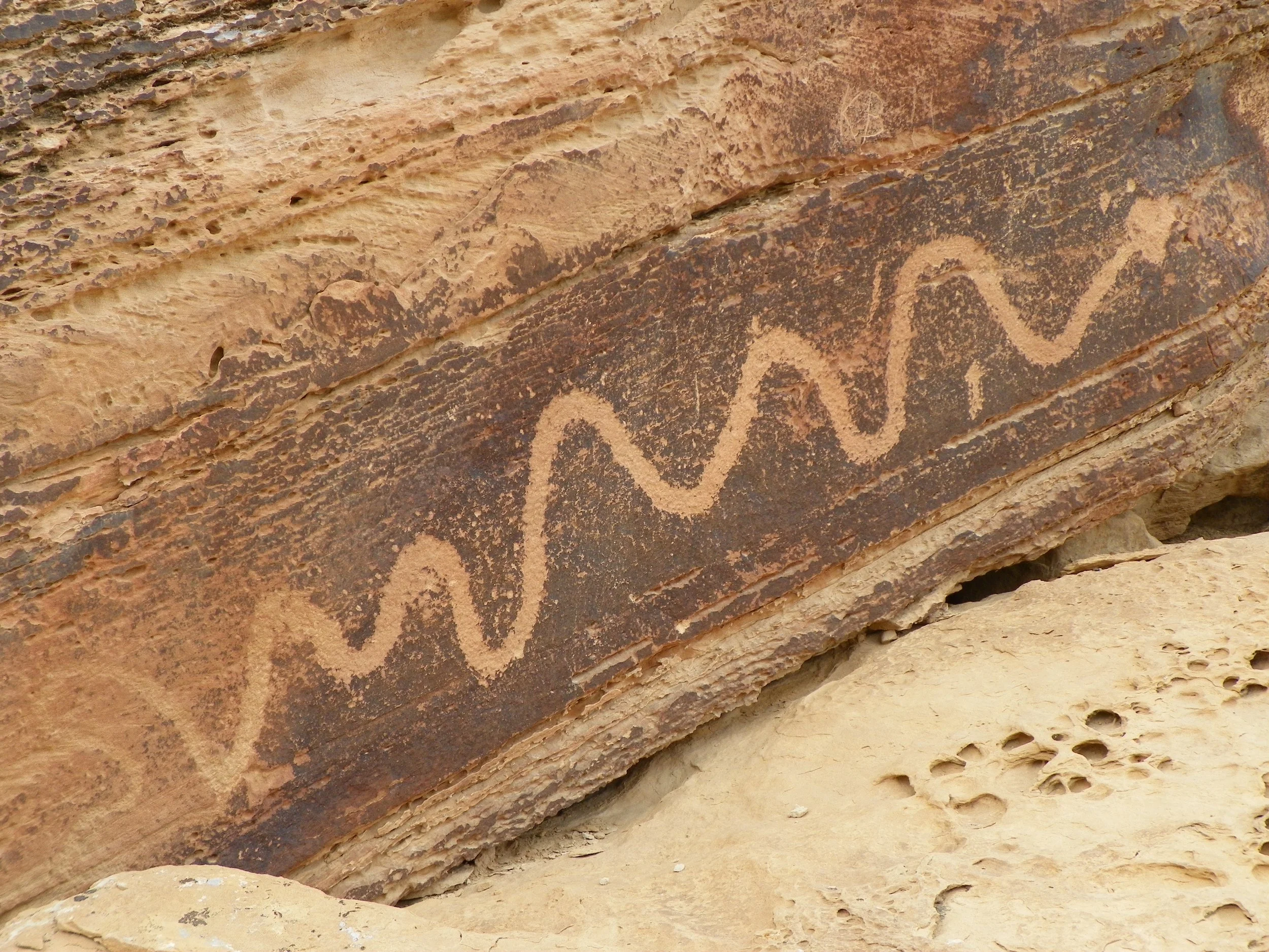 Close-up of ancient petroglyph carving on a reddish-brown stone surface, depicting a snake, with modern names, initials, and scribbles scratched on and around the petroglyph.