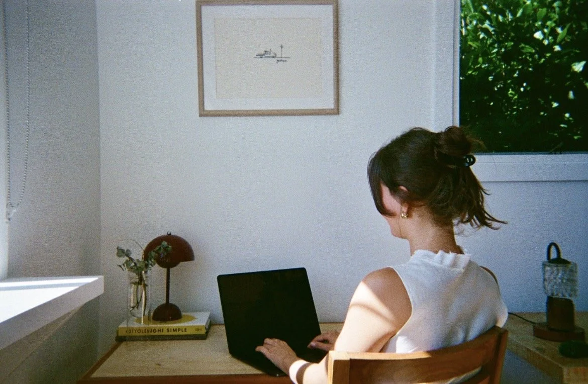 A woman sitting at a wooden desk working on a laptop in a well-lit room with a window showing greenery outside. There is a framed picture on the wall, a small table lamp, and a plant on the desk.