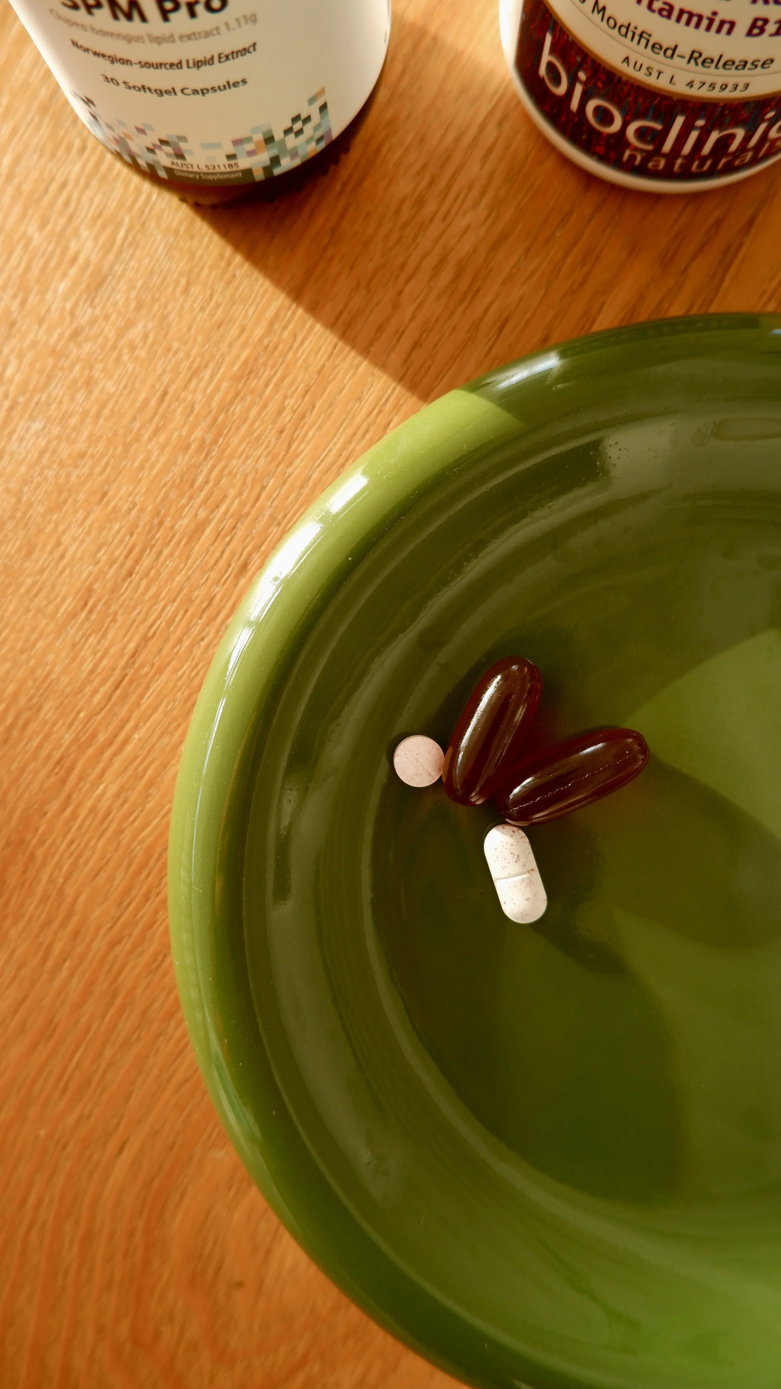 A green ceramic dish with three pills inside, two dark red capsules and one pink and white capsule. Two bottles of supplements are partially visible in the background, on a wooden surface.