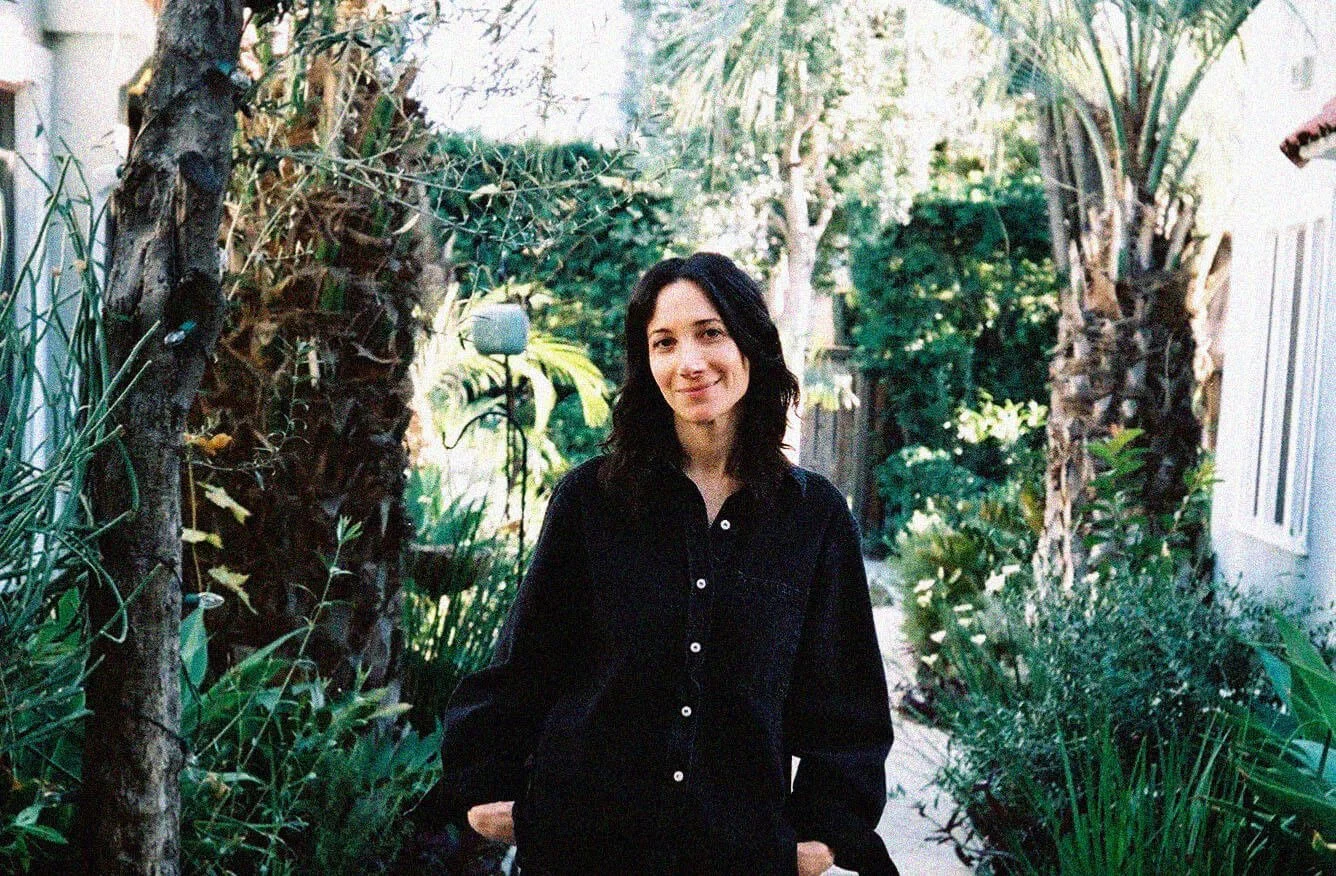 Becky Levi  stands in a lush, green garden with plants and trees, smiling softly at the camera.