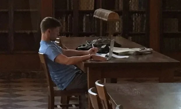 A student studying in UNC's Wilson Library, working on a laptop with books and papers scattered on the desk.