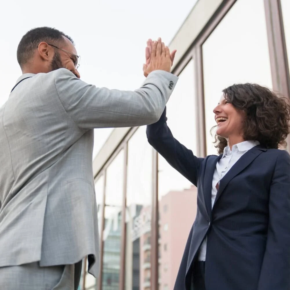 Two business professionals, a man and a woman, smiling and giving each other a high-five outside a modern building.