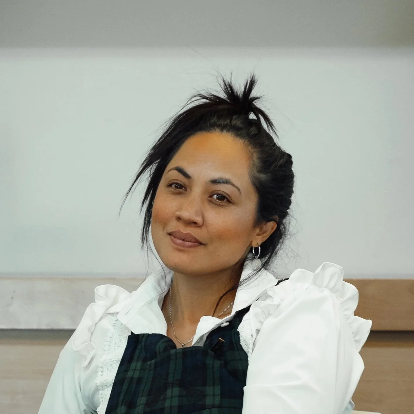 A woman with dark hair in a messy bun, wearing earrings and a white blouse with ruffled sleeves, sitting against a plain light-colored background.