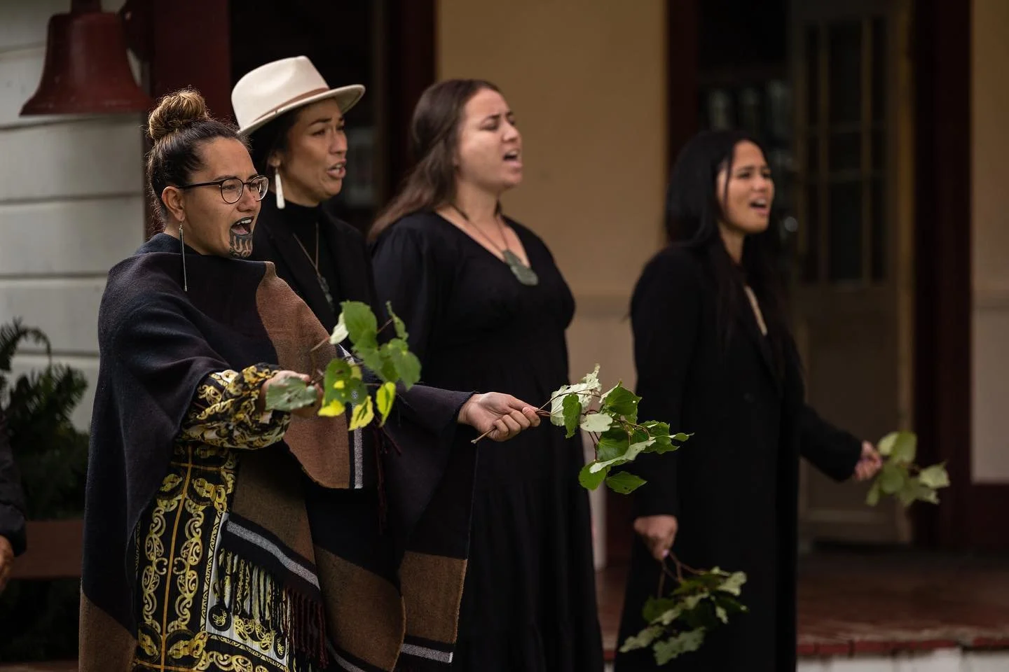 Four women holding branches with green leaves, singing or speaking with emotional expressions indoors.