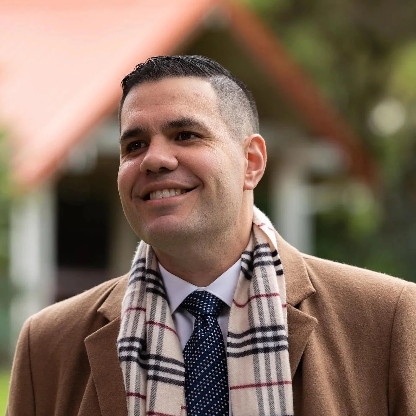 A man outdoors wearing a beige coat, white shirt, dark patterned tie, and a beige plaid scarf, smiling with short dark hair and a well-groomed appearance.