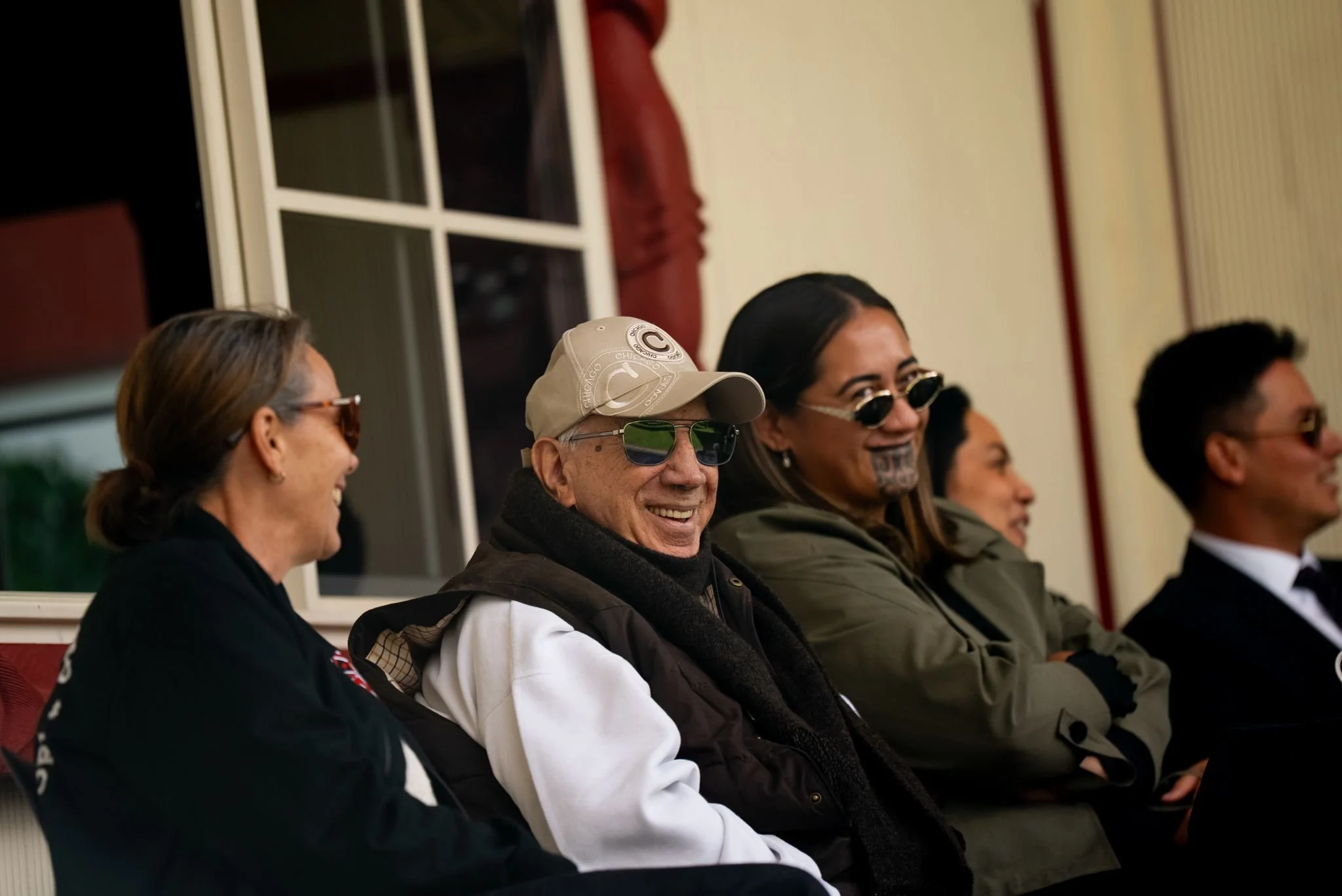 A group of five people sitting side by side in a row, smiling and wearing sunglasses, in an indoor setting with windows and curtains in the background.