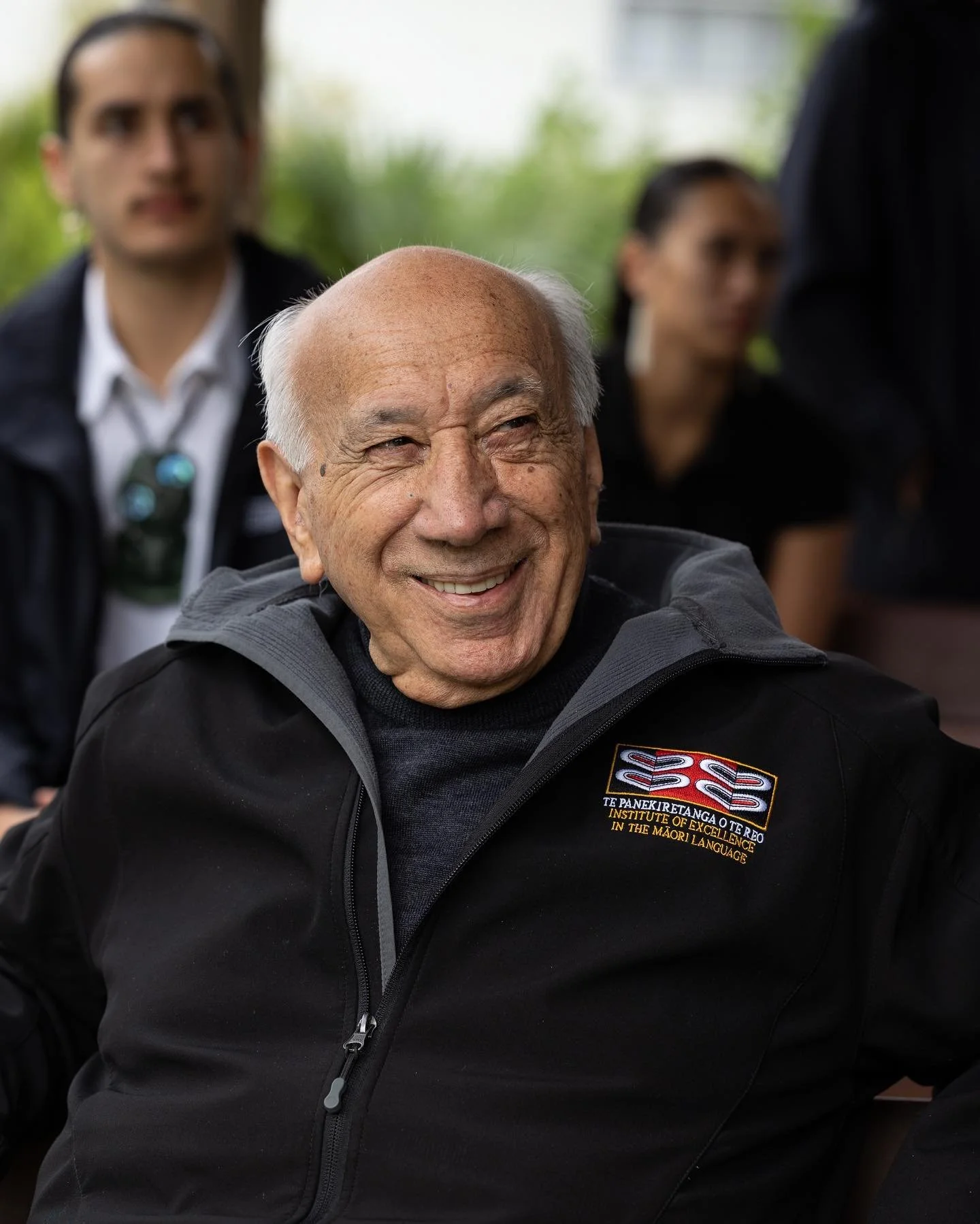 An elderly man smiling while sitting, wearing a black zip-up jacket with an embroidered patch that reads "INSTITUTE OF EXCELLENCE IN THE MAORI LANGUAGE." Two people are blurred in the background, one in a white shirt and sunglasses hanging from his neck, and the other in black with earrings. The setting appears to be outdoors with greenery.